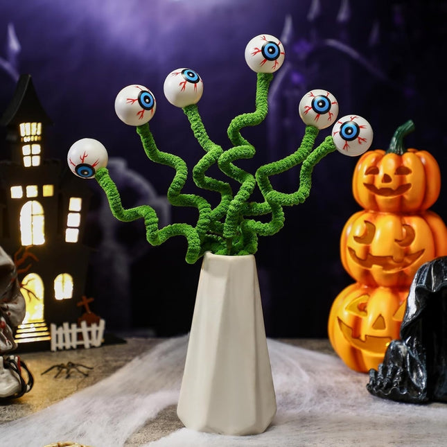 Halloween-themed decoration with eyes on stems in a vase, pumpkins, and a haunted house in the background.